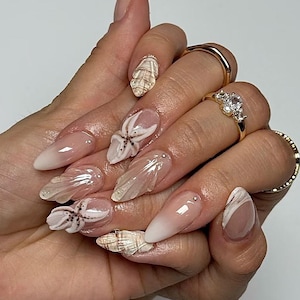 May include: Long, almond-shaped nails with a gradient from nude to white. Some nails feature seashell and starfish designs, while others have a swirl pattern. Gold rings with gemstones adorn the fingers.