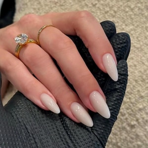 May include: A gold ring with a large round clear stone and a thin gold band with small embedded stones, worn on a finger with long, almond-shaped, glossy light beige nails. The hand is resting against a black textured surface.