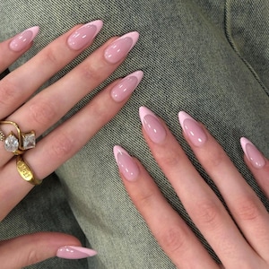 Może przedstawiać: Paznokcie w kształcie migdałów z jasnoróżowym manicure francuskim i błyszczącym wykończeniem. Złoty pierścionek z przezroczystym kamieniem w kształcie gruszki oraz druga złota obrączka z wygrawerowanym napisem 11:11 na palcach.