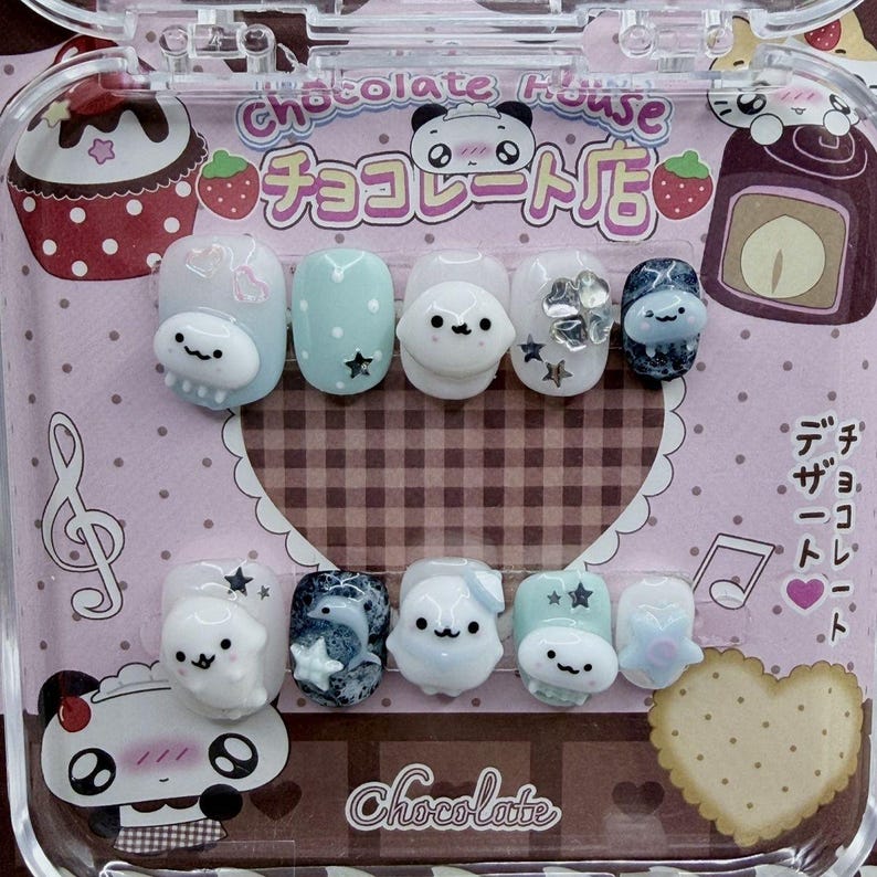 Peut inclure: Un ensemble de faux ongles avec divers motifs, notamment des personnages de dessins anim&eacute;s, des &eacute;toiles et des accents floraux. Les ongles sont dans un &eacute;tui en plastique transparent avec du texte japonais et des illustrations de desserts. La palette de couleurs comprend des pastels et des paillettes.