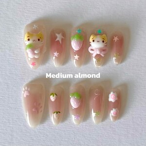 May include: Set of ten medium almond-shaped false nails with a pink and white ombre design. The nails feature various cute designs, including a cat, strawberries, stars, and paw prints.