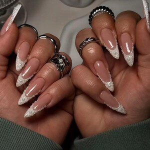 May include: Long, almond-shaped nails featuring a nude base with white French tips adorned with small, raised white pearls along the smile line. Several silver-toned metal rings are worn on the fingers, including textured and smooth bands.