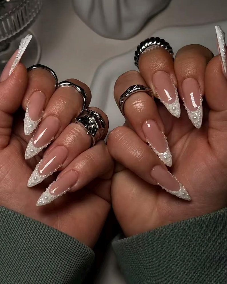 May include: Long, almond-shaped nails featuring a nude base with white French tips adorned with small, raised white pearls along the smile line. Several silver-toned metal rings are worn on the fingers, including textured and smooth bands.