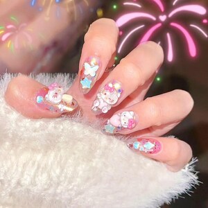 Peut inclure: Une main avec de longs ongles en acrylique transparents d&eacute;cor&eacute;s de personnages de dessins anim&eacute;s en 3D, de papillons et d'&eacute;toiles. Les ongles pr&eacute;sentent des accents roses, bleus et blancs avec des paillettes. La main repose sur une surface blanche duveteuse.