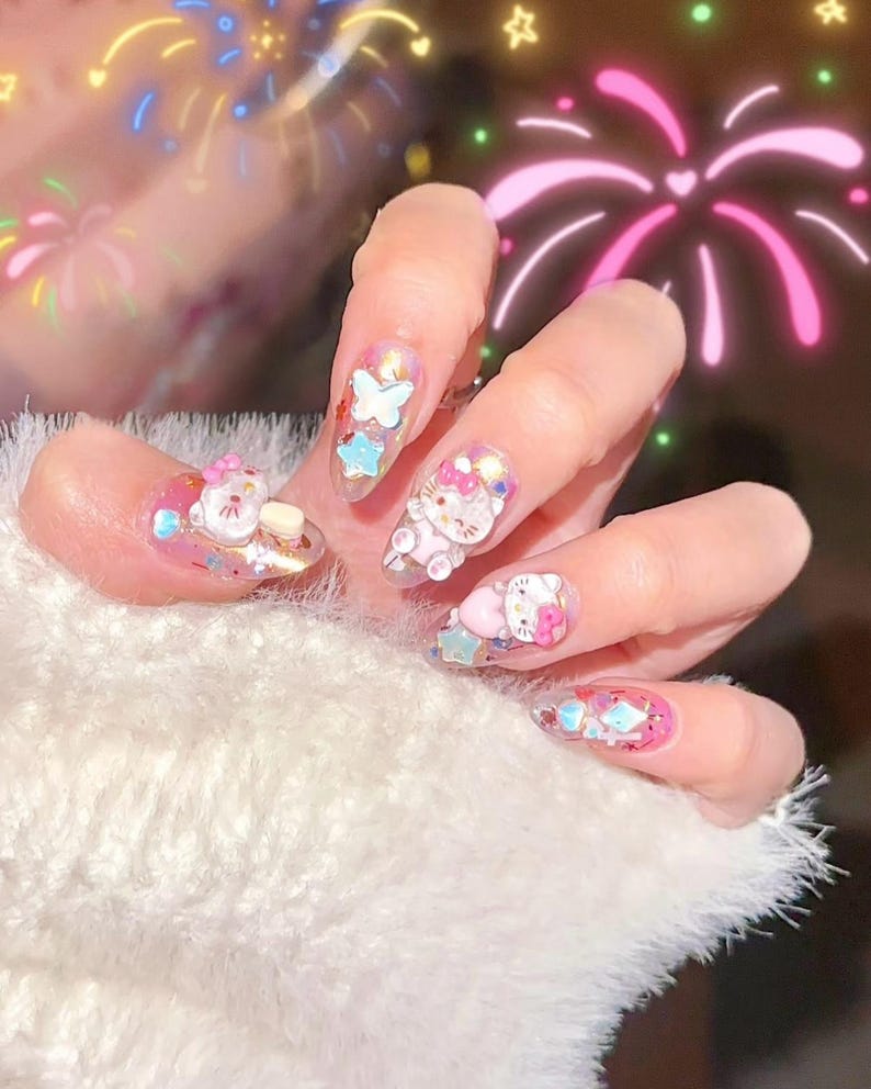 Peut inclure: Une main avec de longs ongles en acrylique transparents d&eacute;cor&eacute;s de personnages de dessins anim&eacute;s en 3D, de papillons et d'&eacute;toiles. Les ongles pr&eacute;sentent des accents roses, bleus et blancs avec des paillettes. La main repose sur une surface blanche duveteuse.