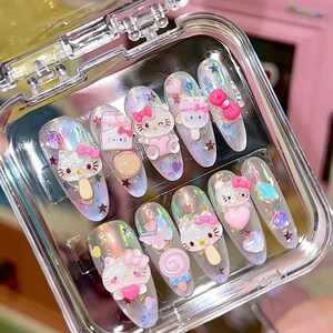 Peut inclure: Un ensemble de dix faux ongles dans un &eacute;tui en plastique transparent. Chaque ongle pr&eacute;sente une base holographique avec divers embellissements 3D, notamment des figurines Hello Kitty, des n&oelig;uds, des c&oelig;urs et des &eacute;toiles. Les ongles sont de formes vari&eacute;es.