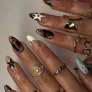 May include: Close-up of manicured nails with various designs and gold rings. Nail art includes tortoiseshell, star, and geometric patterns. Gold rings feature an eye design, stars, and simple bands. The nails are long and almond-shaped.
