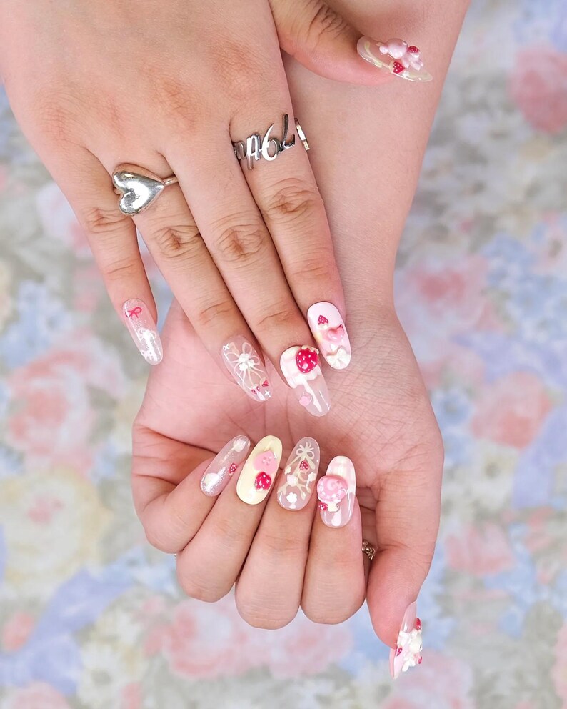 May include: A close-up of a set of long, oval-shaped nails with intricate designs. The nails feature a variety of decorations, including strawberries, bows, and floral patterns. The color palette includes pink, white, and clear tones, creating a delicate and feminine aesthetic.