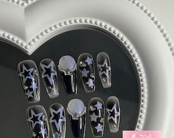 May include: Set of ten black press-on nails with a white star design and silver rhinestones. The nails are displayed on a white heart-shaped background.