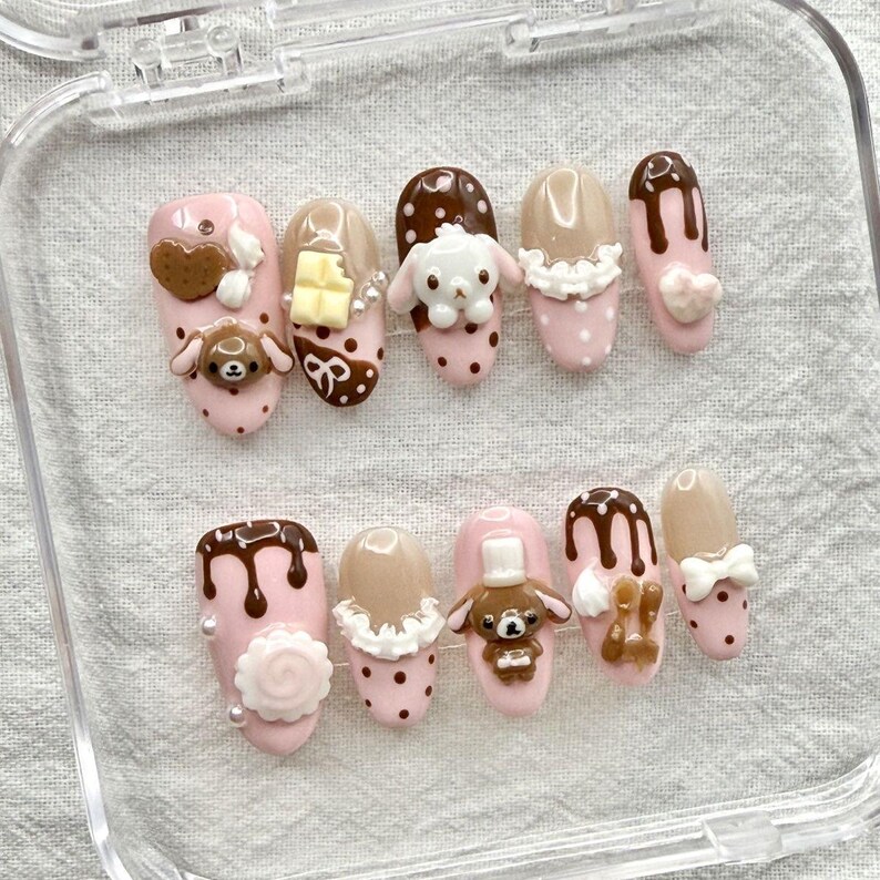 May include: A set of ten artificial nails with a pink base, adorned with various designs. Some nails feature brown chocolate drips, while others have 3D embellishments like a bear, a bunny, and a heart. The nails are in a clear plastic case.