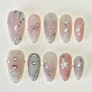 May include: Set of ten pink and white oval-shaped fake nails with silver and white designs. The designs include a bow, a star, a heart, and a spider web.