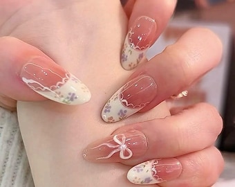 May include: Close-up of a hand with long, oval-shaped nails. The nails feature a transparent base with a pink hue and white tips adorned with a floral pattern. One nail has a white bow design. The overall aesthetic is delicate and feminine.
