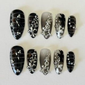 May include: Set of ten black and white press-on nails with silver accents. The nails feature a variety of designs, including a fleur-de-lis, a star, and a cross.