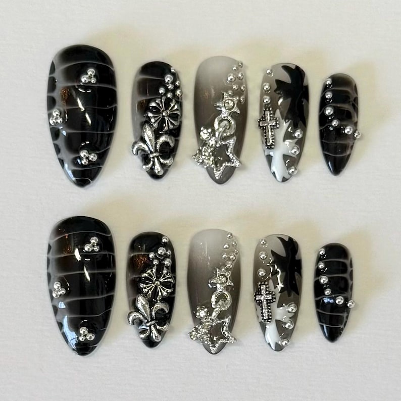 May include: Set of ten black and white press-on nails with silver accents. The nails feature a variety of designs, including a fleur-de-lis, a star, and a cross.