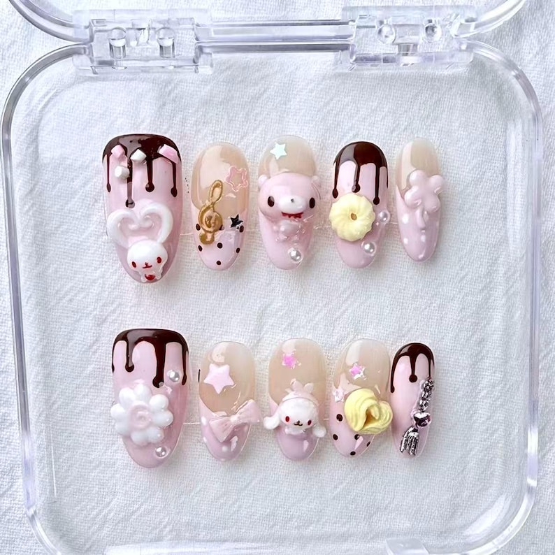 May include: A set of ten press-on nails in a clear plastic case. Each nail is painted light pink and decorated with various 3D designs, including chocolate drips, flowers, bows, and cartoon characters. Some nails have pearl accents.