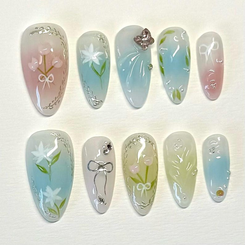 Pode incluir: Dez unhas posti&ccedil;as longas e ovais com um degrad&ecirc; de azul claro, rosa ou amarelo. Cada unha &eacute; decorada com um design floral diferente, incluindo flores, folhas e uma borboleta.