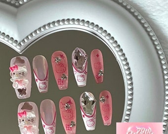 May include: Pink and white press-on nails with Hello Kitty designs, rhinestones, and a pink heart accent. The nails are displayed on a white heart-shaped mirror with the text "79th Street Press On Nails".