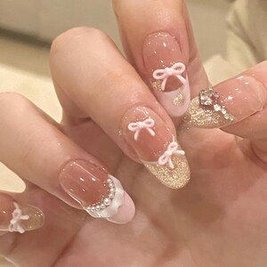 K&ouml;nnte beinhalten: Nahaufnahme einer Hand mit rosa und wei&szlig;er Nagelkunst. Die N&auml;gel sind mit kleinen rosa Schleifen und Glitzer verziert. Einige N&auml;gel haben eine wei&szlig;e Spitze mit einem perlmuttartigen Design.