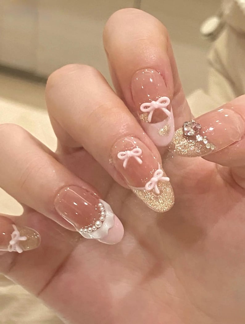 K&ouml;nnte beinhalten: Nahaufnahme einer Hand mit rosa und wei&szlig;er Nagelkunst. Die N&auml;gel sind mit kleinen rosa Schleifen und Glitzer verziert. Einige N&auml;gel haben eine wei&szlig;e Spitze mit einem perlmuttartigen Design.