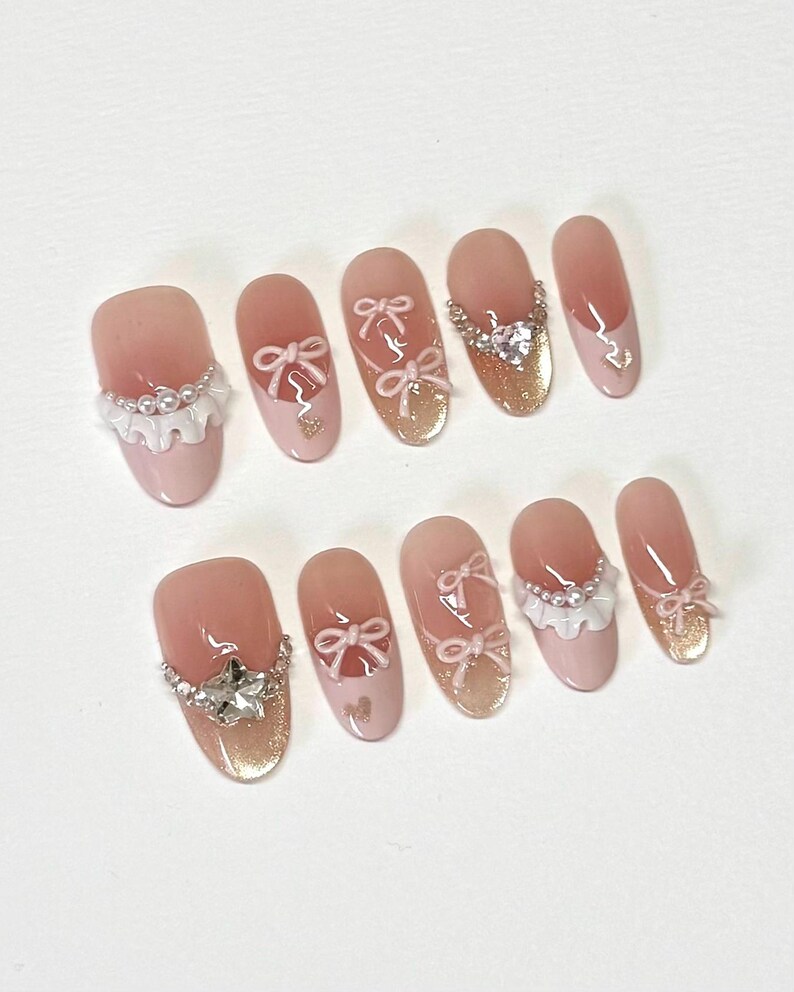 K&ouml;nnte beinhalten: Set aus zehn ovalen, falschen N&auml;geln in Rosa und Wei&szlig; mit verschiedenen Verzierungen, darunter Schleifen, Strasssteine und Perlenakzente.