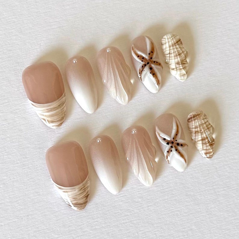 May include: A set of ten artificial nails with a beach theme. The nails feature a variety of designs, including a light brown base with a swirl pattern, a gradient from light brown to white, and a starfish design. Some nails have small, clear embellishments.