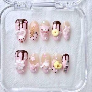 May include: A set of ten artificial nails in a clear plastic case. Each nail is painted light pink and decorated with various designs, including chocolate drips, cartoon characters, bows, stars, and flowers. Some nails have pearl accents.