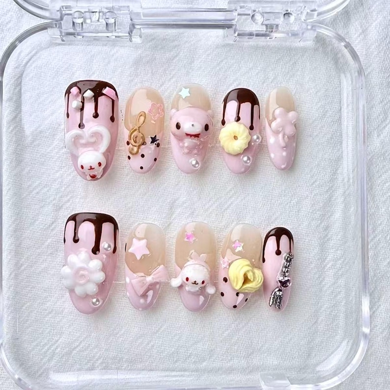 May include: A set of ten artificial nails in a clear plastic case. Each nail is painted light pink and decorated with various designs, including chocolate drips, cartoon characters, bows, stars, and flowers. Some nails have pearl accents.