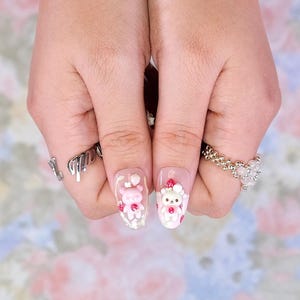 May include: Oval-shaped acrylic nails with a clear base, featuring 3D pink teddy bear and strawberry designs. The nails are adorned with small red strawberries and white accents. Silver rings are worn on the fingers.