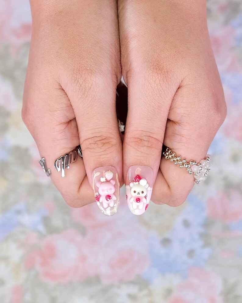 May include: Oval-shaped acrylic nails with a clear base, featuring 3D pink teddy bear and strawberry designs. The nails are adorned with small red strawberries and white accents. Silver rings are worn on the fingers.