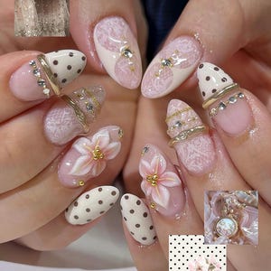 May include: Manicured nails featuring a mix of designs including white lace patterns, brown polka dots on a cream base, and three-dimensional pink floral accents with gold beads. The nails are adorned with gold rings, small clear crystals, and thin gold chains.
