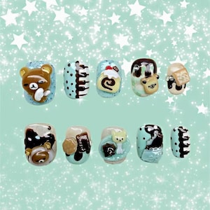 May include: A collection of ten artificial nails, each decorated with a different design. The nails feature cartoon bears, cake rolls, and other dessert-themed decorations. The base colours include light blue, brown, and white, with black polka dots and accents. The nails are set against a light green background with white star shapes.