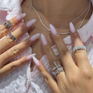 Può includere: Una collezione di anelli argentati indossati su dita con unghie lunghe a mandorla color rosa chiaro. I gioielli includono un anello con pietre trasparenti, un anello a forma di croce e due anelli con lettere. Una sottile catenina d'argento su una camicia rosa.
