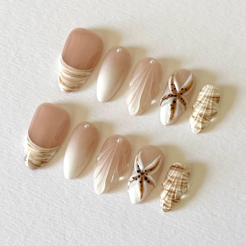 May include: A set of ten artificial nails with a beach theme. The nails feature a variety of designs, including a marbled effect, a gradient, a seashell design, and a starfish design. The color palette includes shades of brown, white, and beige.