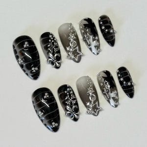 May include: Set of ten black and white false nails with silver accents. The nails have a gradient design with black on one side and white on the other. Each nail features a different silver design, including a star, a cross, and a fleur-de-lis.