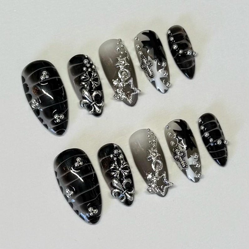 May include: Set of ten black and white false nails with silver accents. The nails have a gradient design with black on one side and white on the other. Each nail features a different silver design, including a star, a cross, and a fleur-de-lis.