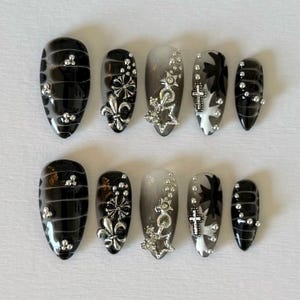 May include: Set of ten black and silver press-on nails with various designs. The nails have a black base with silver accents, including crosses, stars, and fleur-de-lis. Some nails have a gradient effect, transitioning from black to silver.