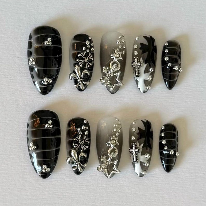May include: Set of ten black and silver press-on nails with various designs. The nails have a black base with silver accents, including crosses, stars, and fleur-de-lis. Some nails have a gradient effect, transitioning from black to silver.