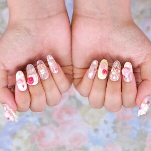 May include: A set of long, almond-shaped acrylic nails. Each nail features a unique design, including strawberries, bows, and floral patterns. The color palette includes white, pink, and clear accents, creating a whimsical and feminine aesthetic.