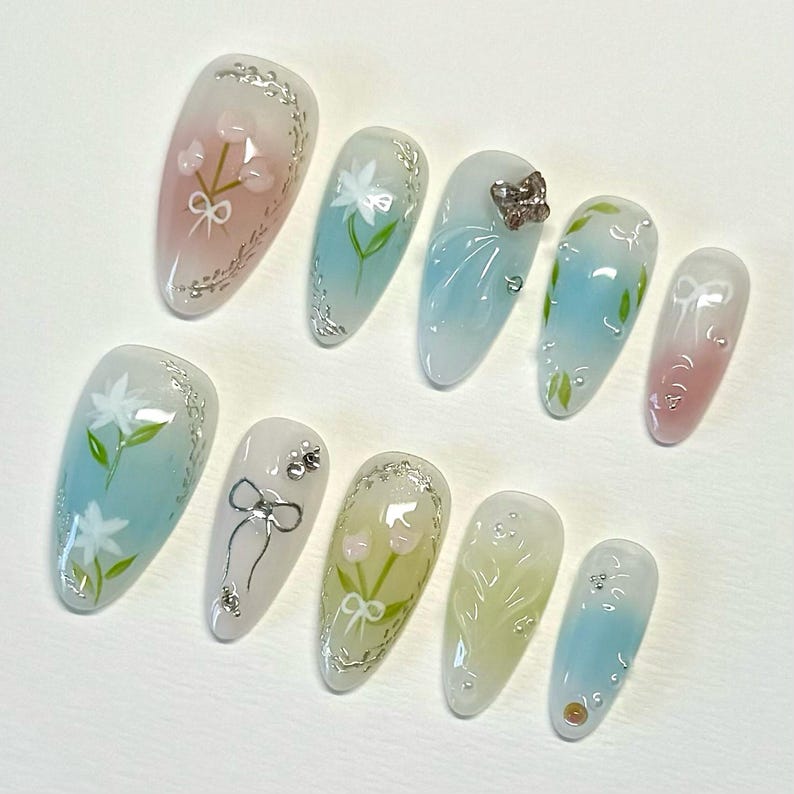 Pode incluir: Dez unhas posti&ccedil;as longas e ovais com base transparente e desenhos pintados. Os desenhos incluem flores, borboletas e la&ccedil;os em tons de rosa, azul e verde.