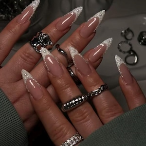May include: Long, stiletto-shaped nails featuring a nude base color with white French tips accented by small, raised white pearls along the smile line. Several silver-toned metal rings are worn on the fingers, including a chunky band and a chain link style.