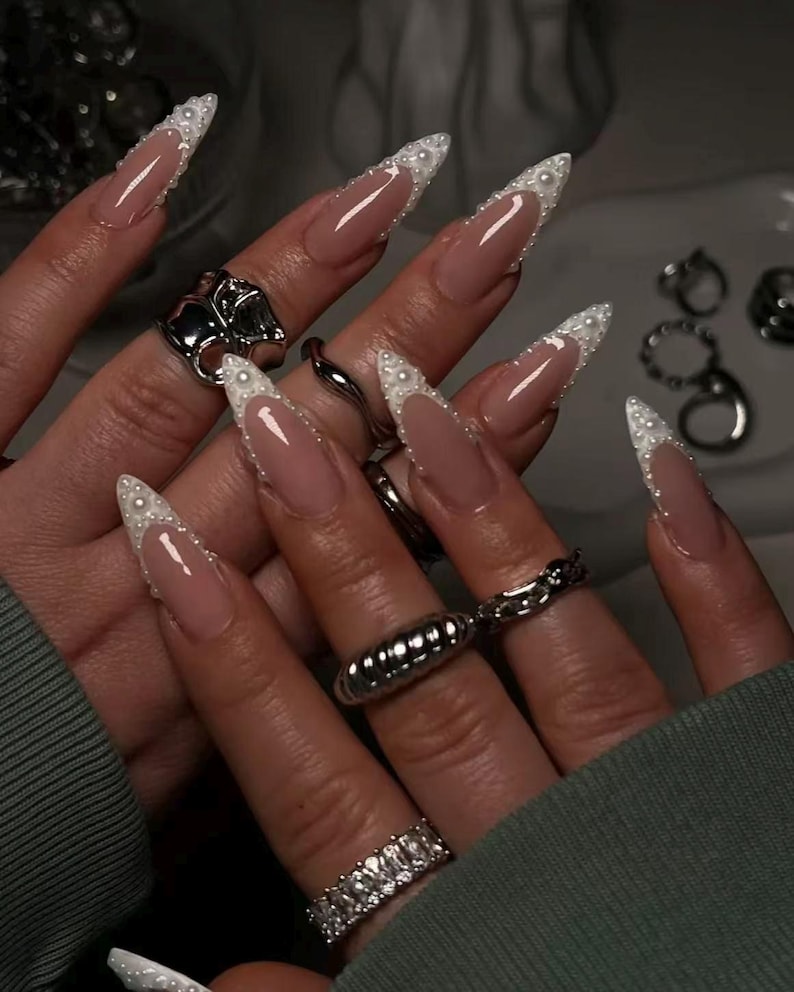May include: Long, stiletto-shaped nails featuring a nude base color with white French tips accented by small, raised white pearls along the smile line. Several silver-toned metal rings are worn on the fingers, including a chunky band and a chain link style.