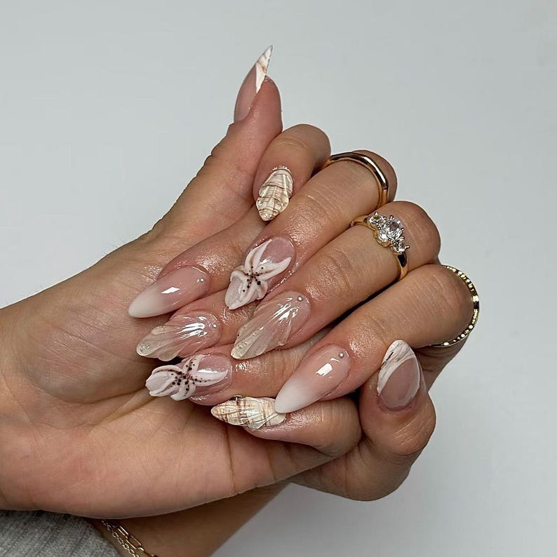 May include: Long, almond-shaped nails with a gradient from nude to white. Some nails feature seashell and starfish designs, while others have a swirl pattern. Gold rings with gemstones adorn the fingers.
