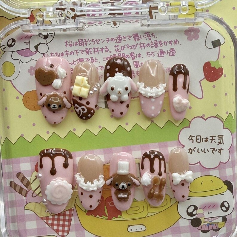 May include: A set of press-on nails in a clear case. The nails are primarily pink, with various designs including cartoon characters, chocolate, and polka dots. Some nails have 3D embellishments. The text "Today is a nice day" is visible.