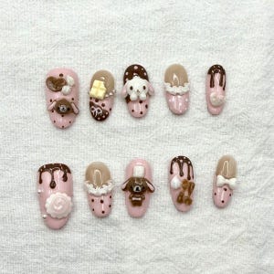 May include: A set of ten pink artificial nails with various 3D designs. The designs include chocolate drips, teddy bears, bunnies, and bows. Some nails have white polka dots and a rose.