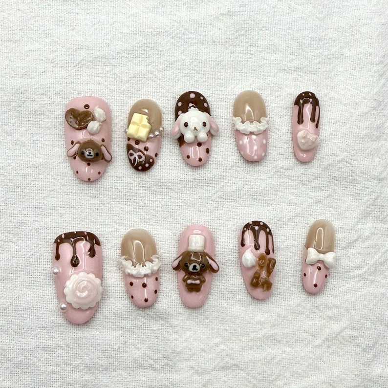 May include: A set of ten pink artificial nails with various 3D designs. The designs include chocolate drips, teddy bears, bunnies, and bows. Some nails have white polka dots and a rose.