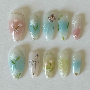 Pode incluir: Conjunto de dez unhas posti&ccedil;as com um design degrad&ecirc; de azul claro e rosa. Cada unha apresenta um design floral diferente, incluindo uma borboleta, uma flor e um la&ccedil;o.