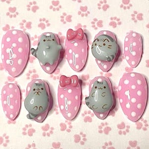 May include: A set of ten press-on nails. Each nail is pink with white polka dots. Some nails feature a gray cat design, while others have a pink bow. The nails are oval-shaped.