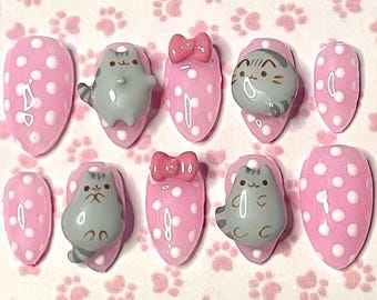 May include: A set of ten press-on nails. Each nail is pink with white polka dots. Some nails feature a gray cat design, while others have a pink bow. The nails are oval-shaped.