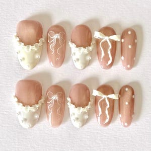 May include: A set of ten artificial nails in a variety of designs. The nails feature a light pink base with white accents, including bows, polka dots, and a ruffled edge with small pearl-like embellishments.