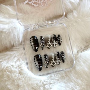 May include: A set of black and white press-on nails with silver accents. The nails feature a cross and floral design.